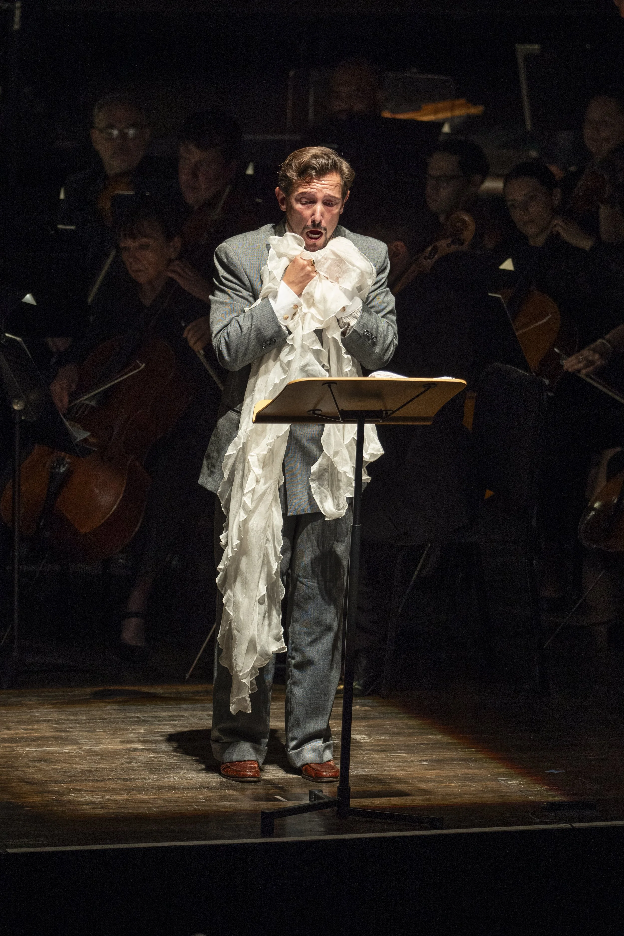 A man in a suit singing on stage with an orchestra in the background, holding a scarf.