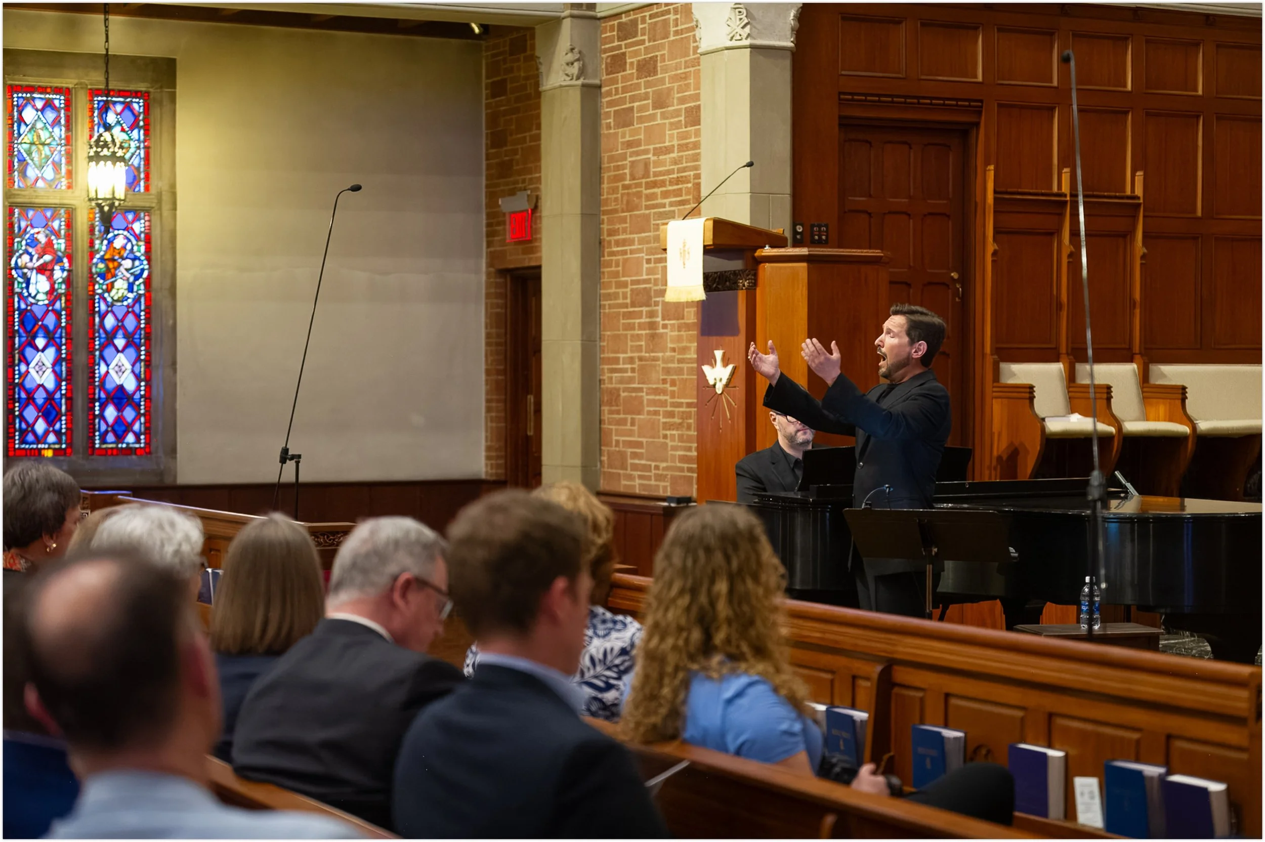 Aaron Blake singing in a sanctuary, standing next to a grand piano, with a congregation seated in pews in front of him, inside a church with stained glass windows.