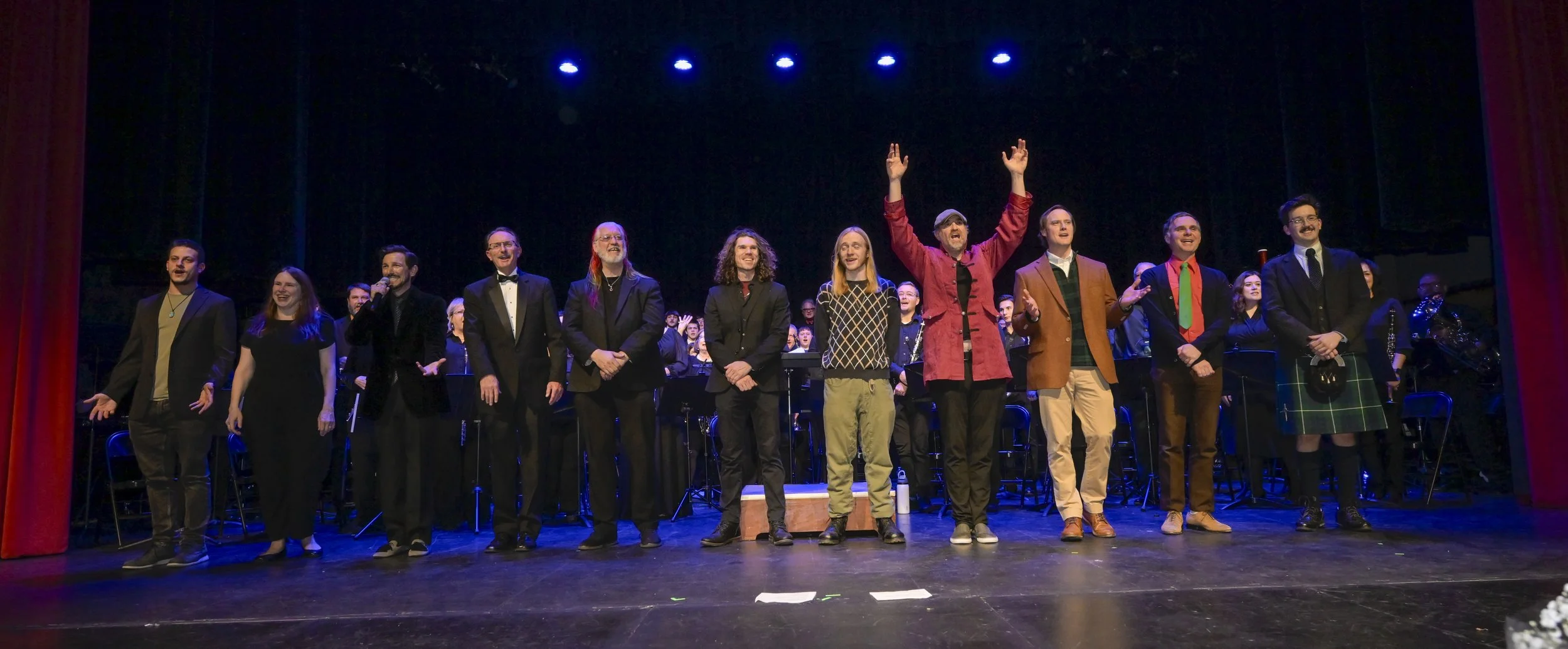 Group of performers standing on stage, some waving, with a choir or orchestra behind them, in a theater with red curtains.