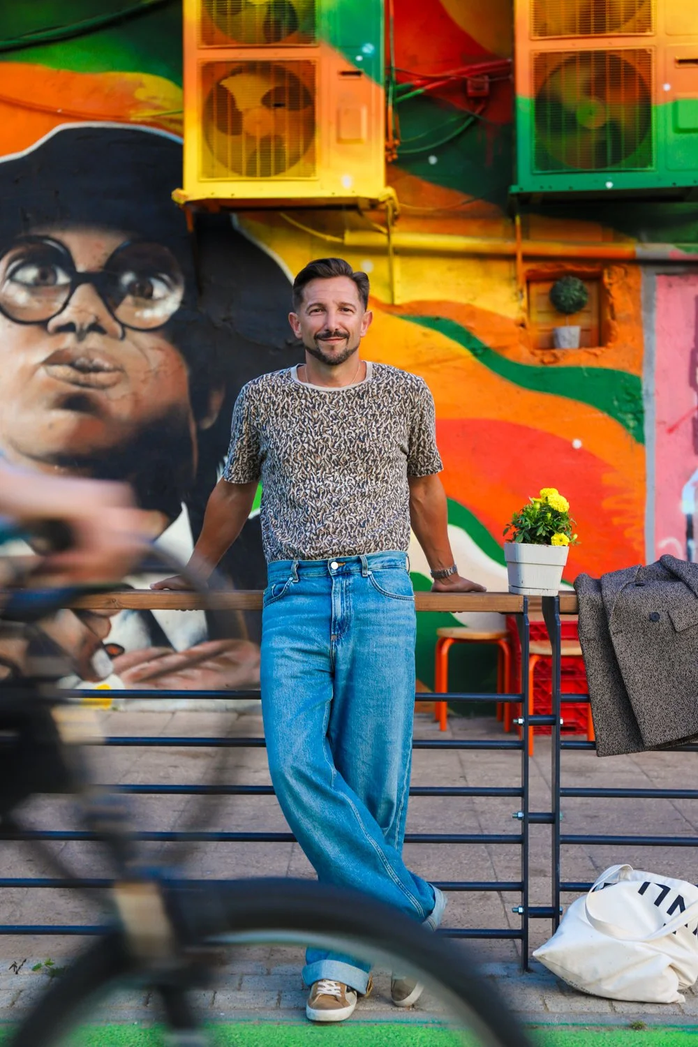 Aaron Blake in jeans and a tshirt leans against a metal fence in front of a colorful mural