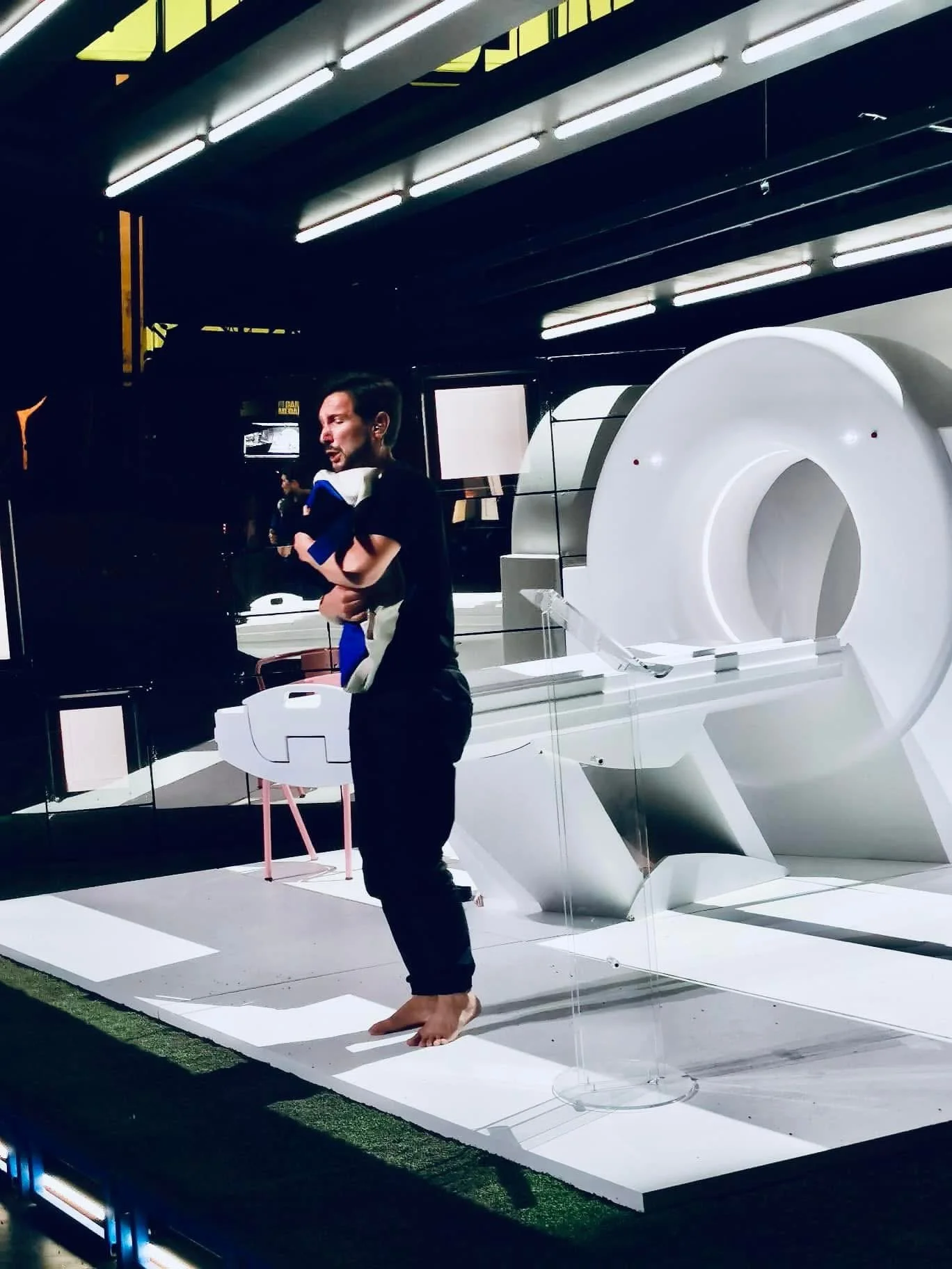 Aaron Blake standing on a white stage inside a mirrored set with an MRI scanner in the background.