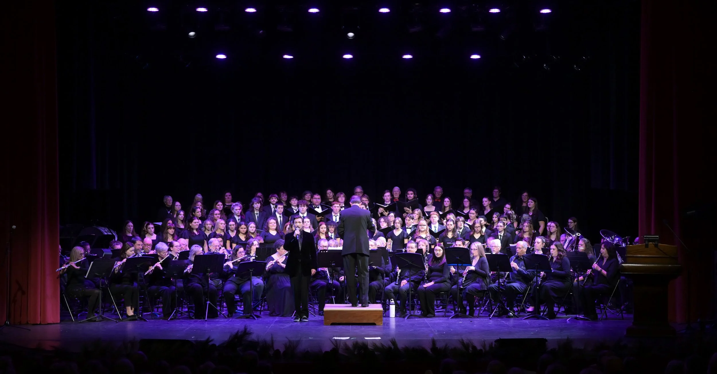 A large choir and orchestra performing on stage with a conductor and a solo singer under purple stage lighting.
