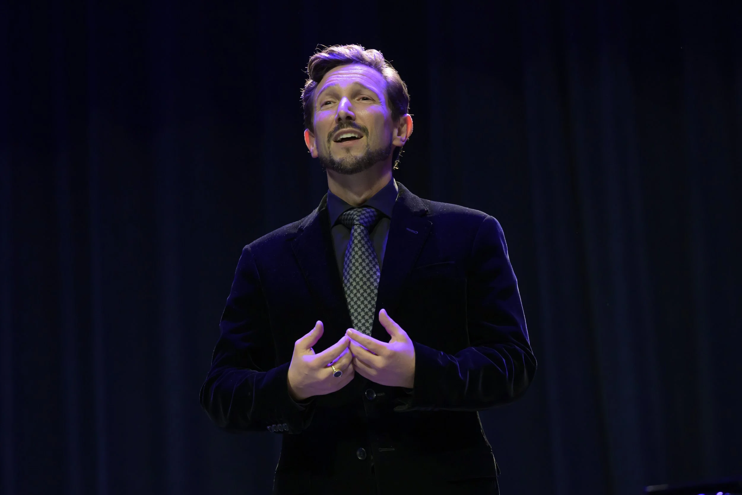 Aaron Blake wearing a black suit and patterned tie, singing on a stage with dark curtains behind him.