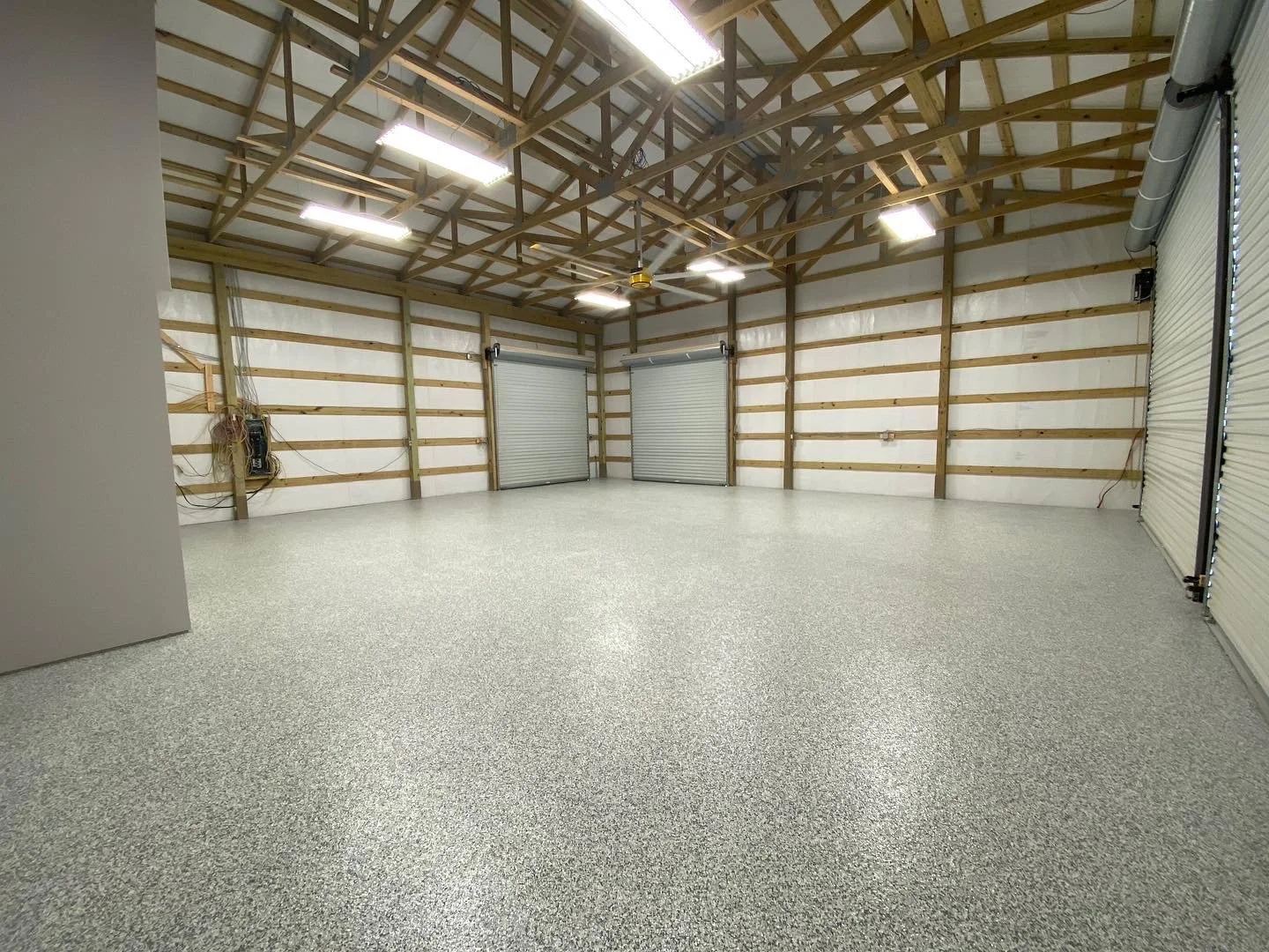 Empty garage with epoxy-coated floor, unfinished wood framing, two small garage doors, and industrial lighting.