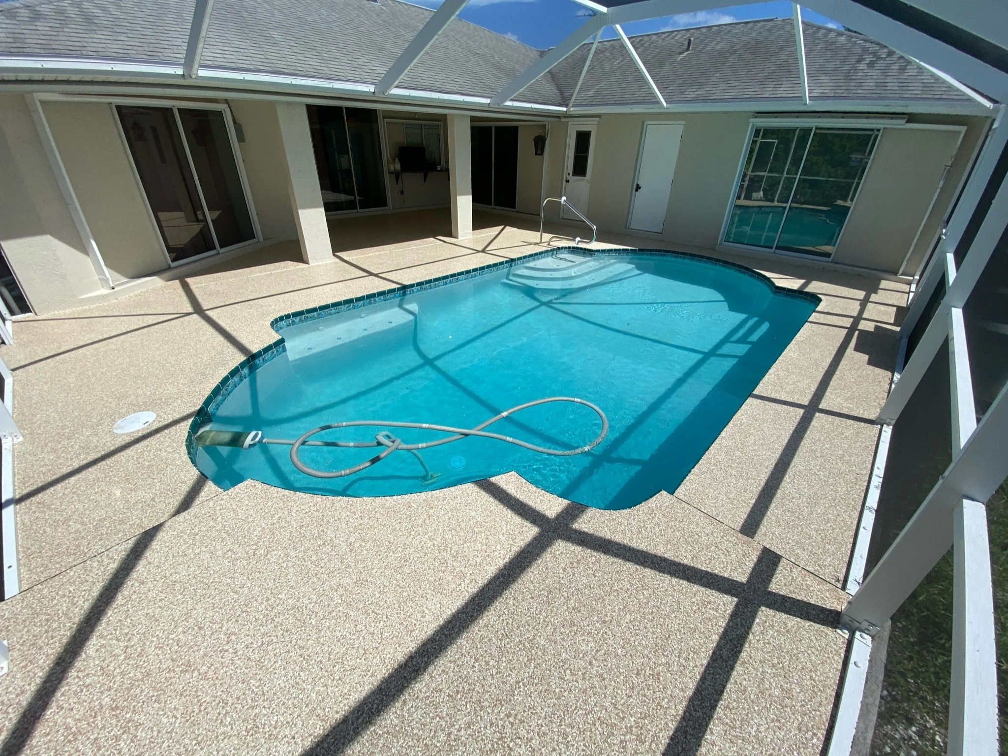 Empty backyard pool enclosed with screened porch, with pool cleaning equipment floating on the water and shadows cast by the screen structure.