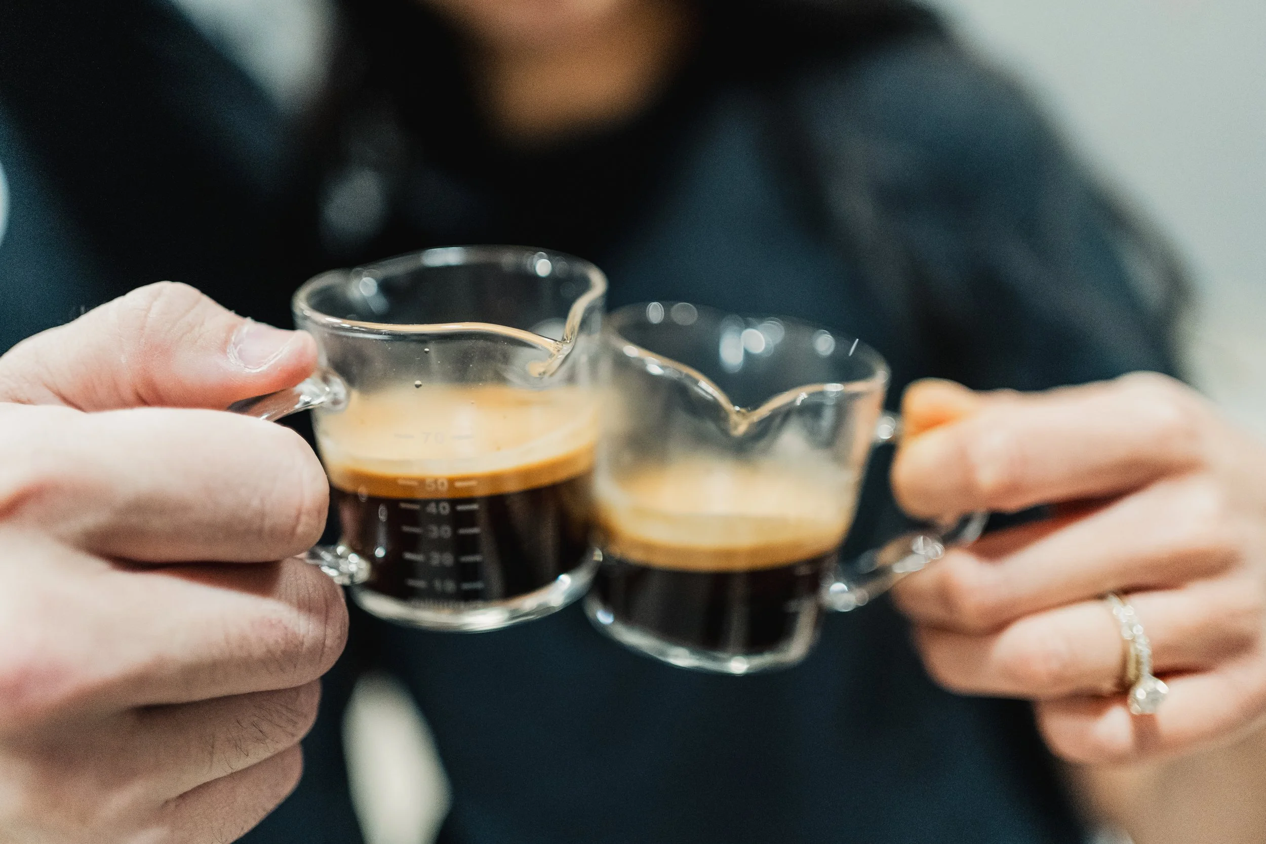 A person is holding two small glass cups of espresso coffee in each hand, with a black shirt and a ring visible on one hand.