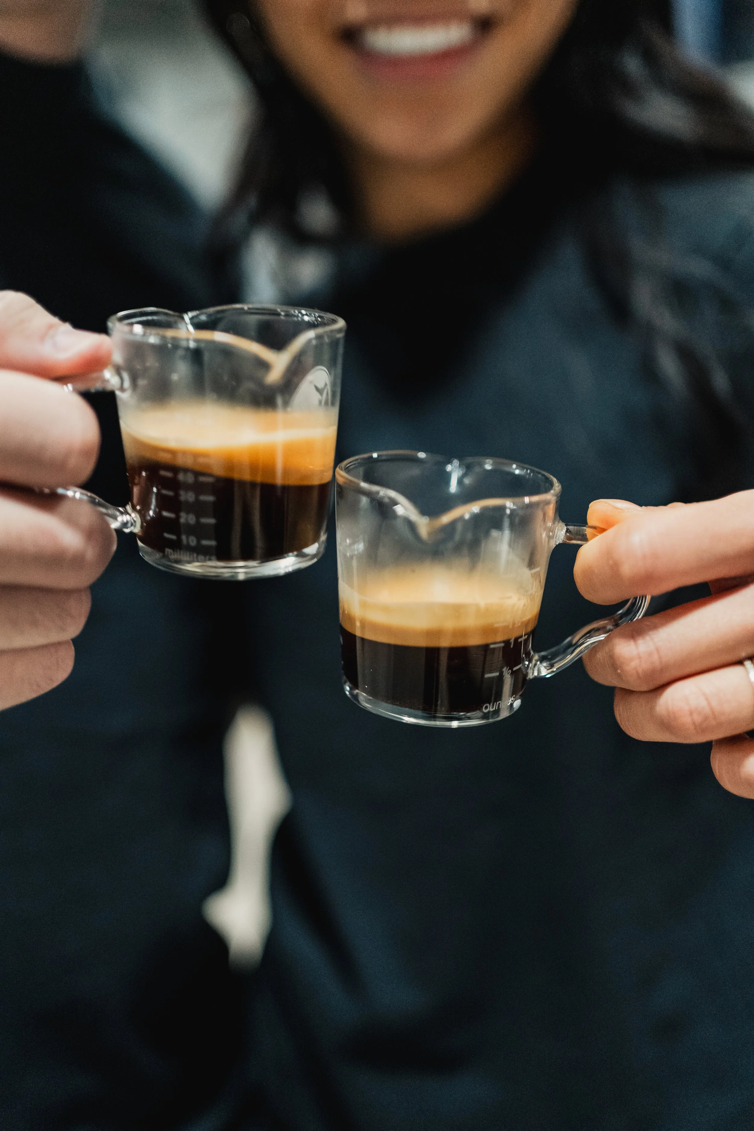 Person holding two small glass cups of espresso, smiling.