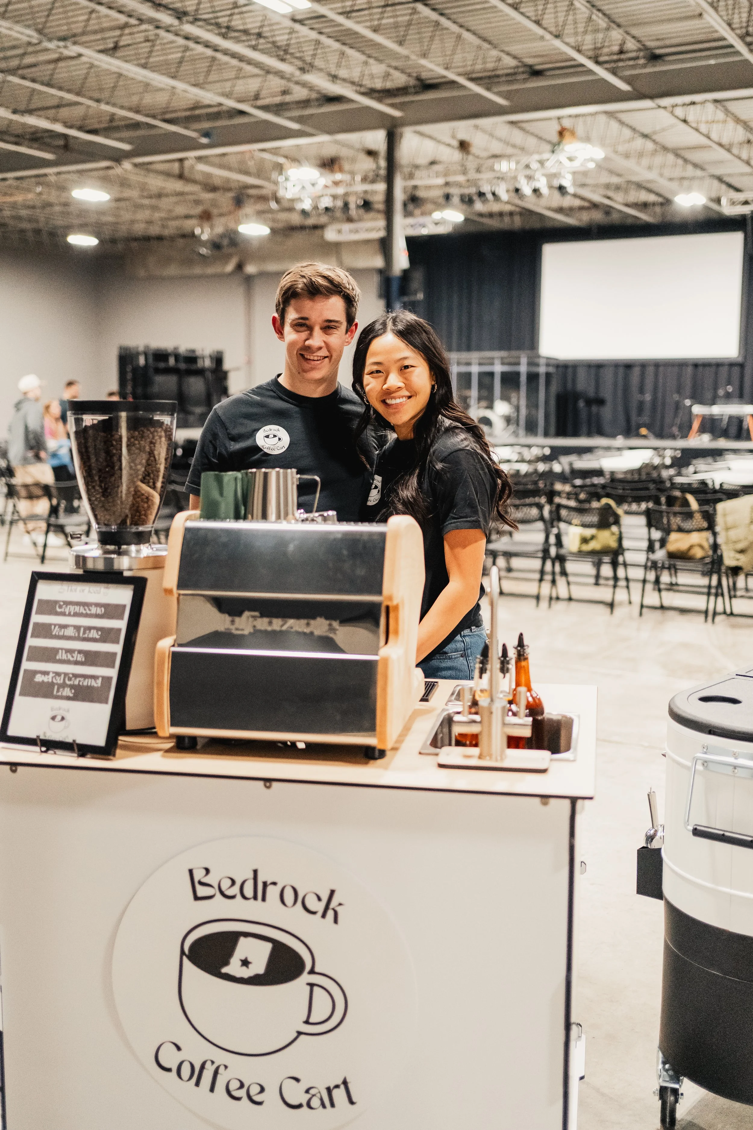 Two smiling people standing behind a coffee cart in a large indoor event space.
