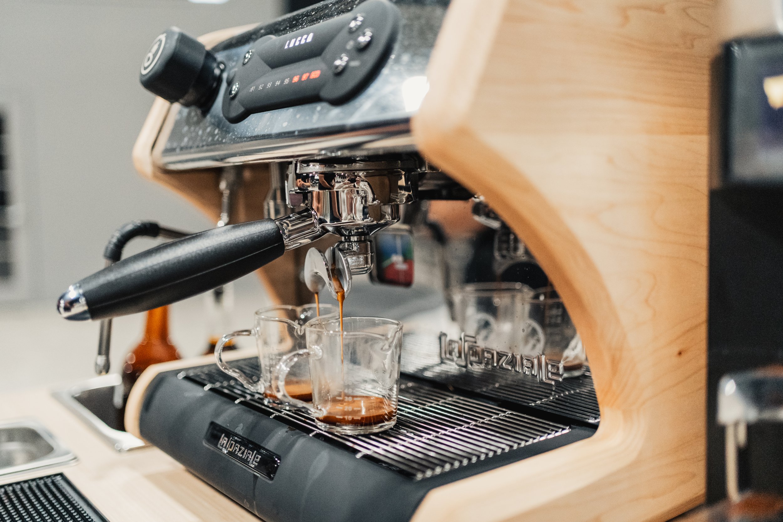 A close-up view of a espresso machine pouring coffee into glass cups, with a wooden frame and a stainless steel drip tray.