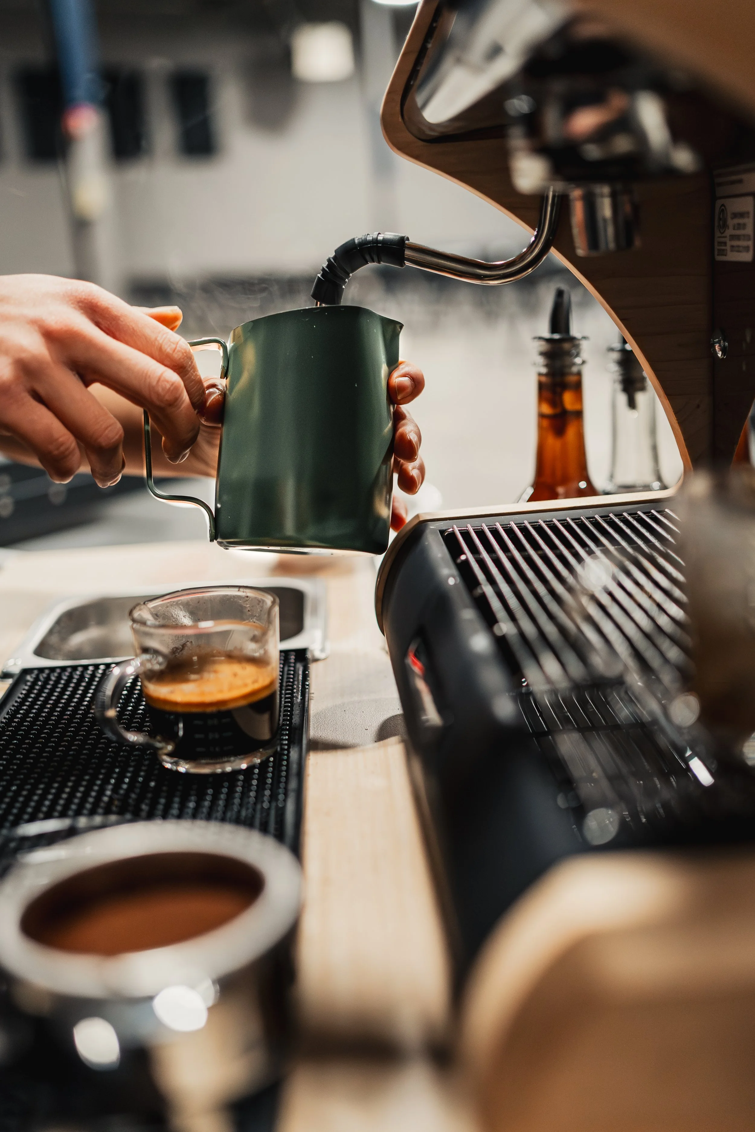 Person pouring steam from a green milk frothing pitcher into a glass cup with espresso, on a coffee station with a coffee machine and accessories.