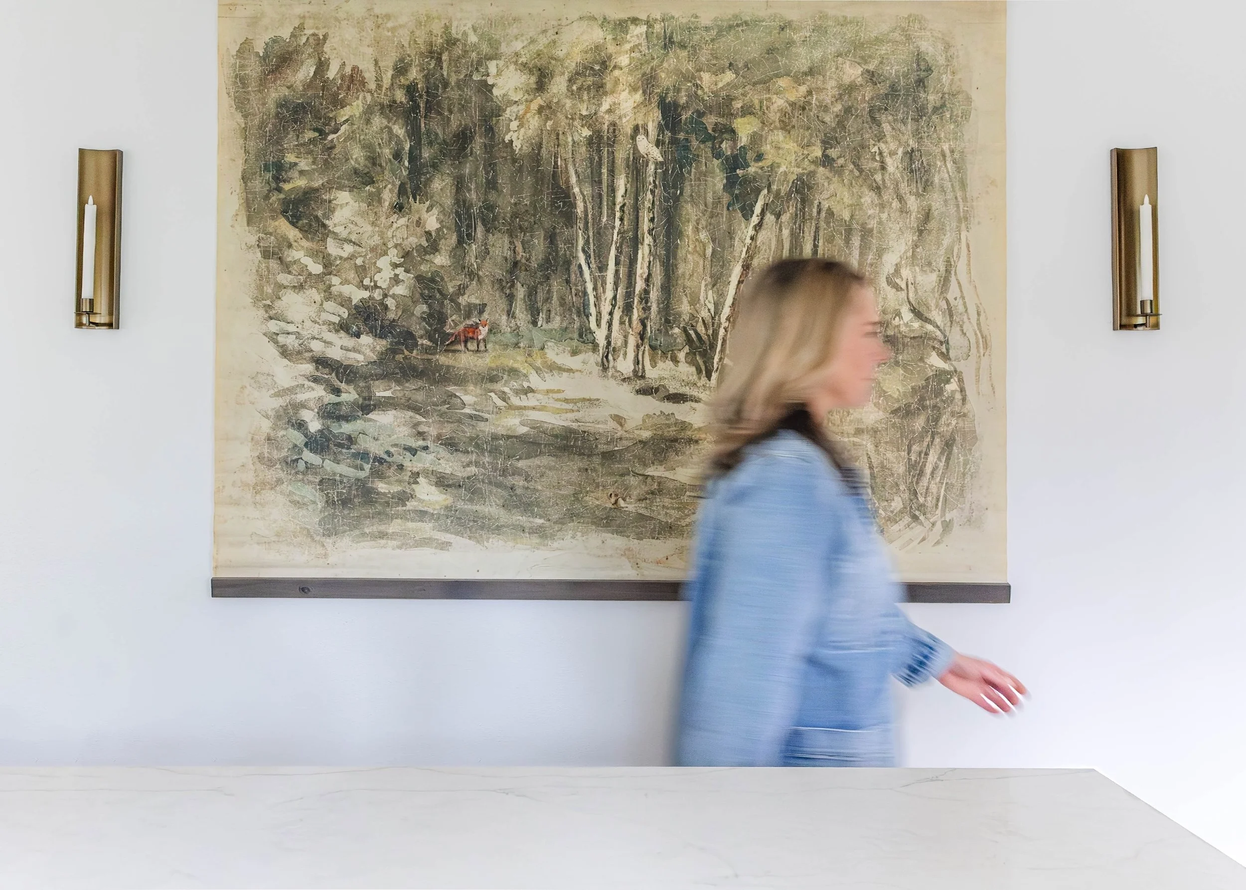 A woman with blonde hair, dressed in a blue jacket, walking past a large landscape painting of a forest scene with trees and a fox. The painting is mounted on a white wall flanked by two wall sconces with candles.