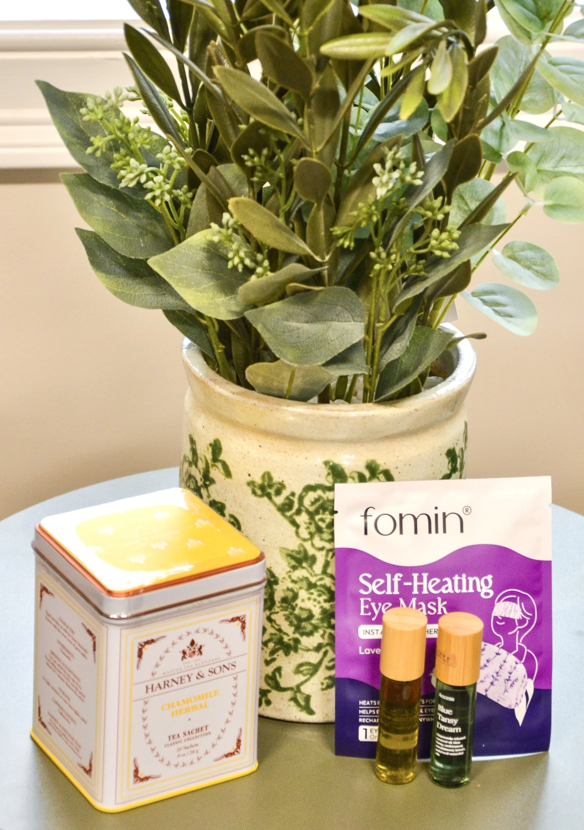 A potted plant with green leaves, two essential oil bottles, a box of Harney & Sons Chamomile herbal tea sachets, and a packet of Fomin Self-Healing Eye Mask on a table.