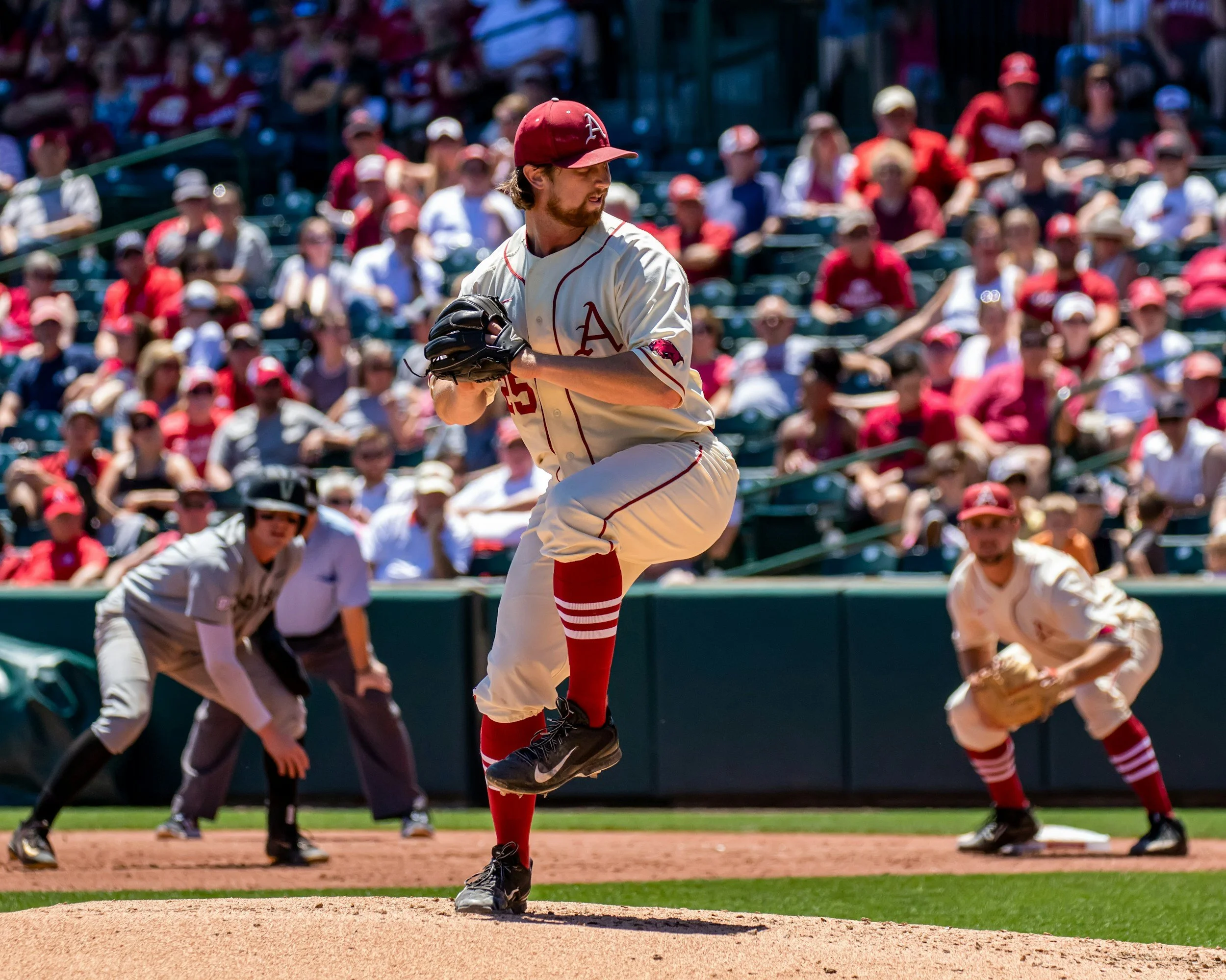 A baseball player wearing a white uniform with red accents, a red cap with an 'A' logo, and red socks is jumping on the field, with a crowd of spectators in the background.