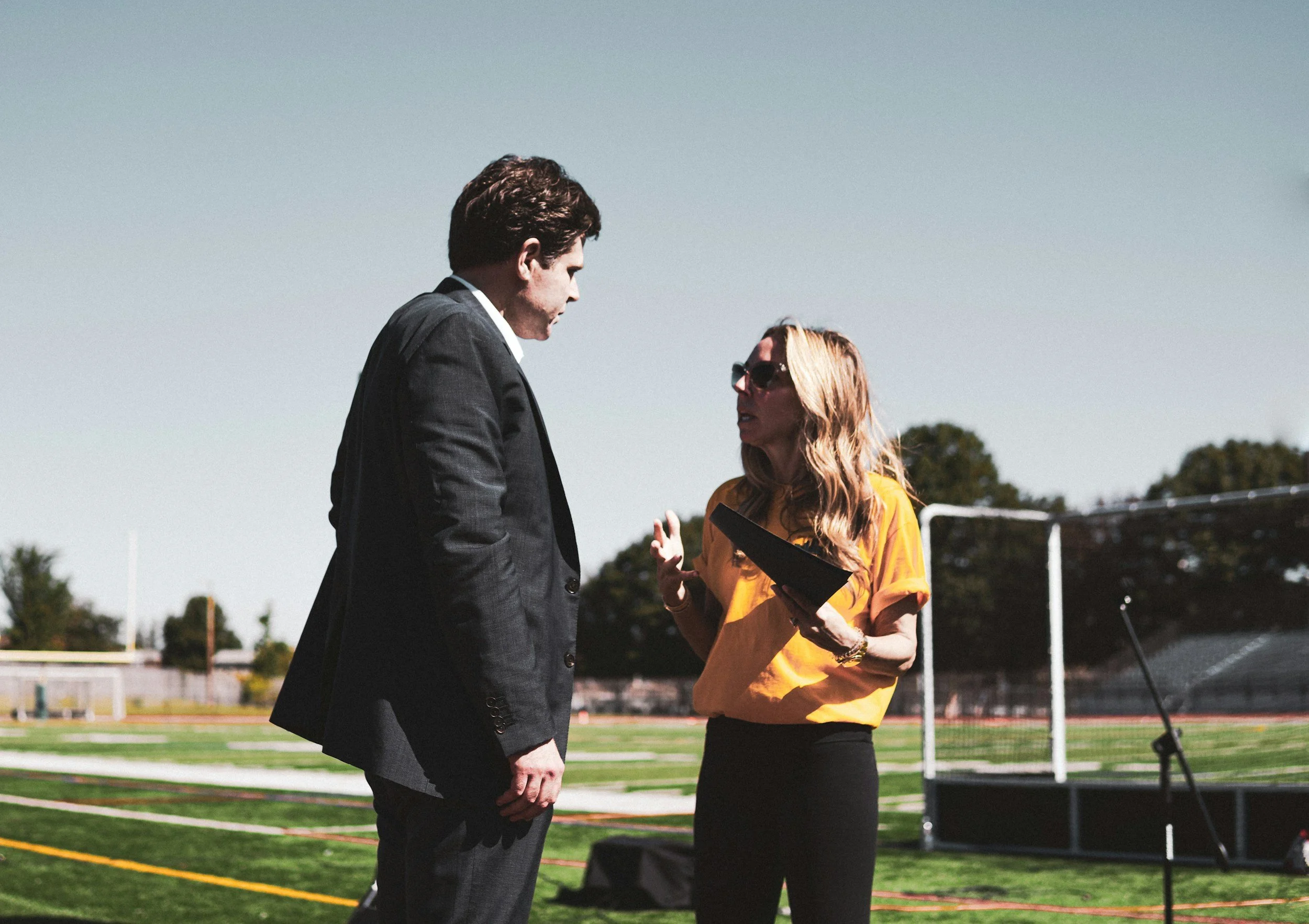 A man in a suit and a woman in sunglasses are having a conversation outdoors on a track field, with trees and sports equipment in the background.
