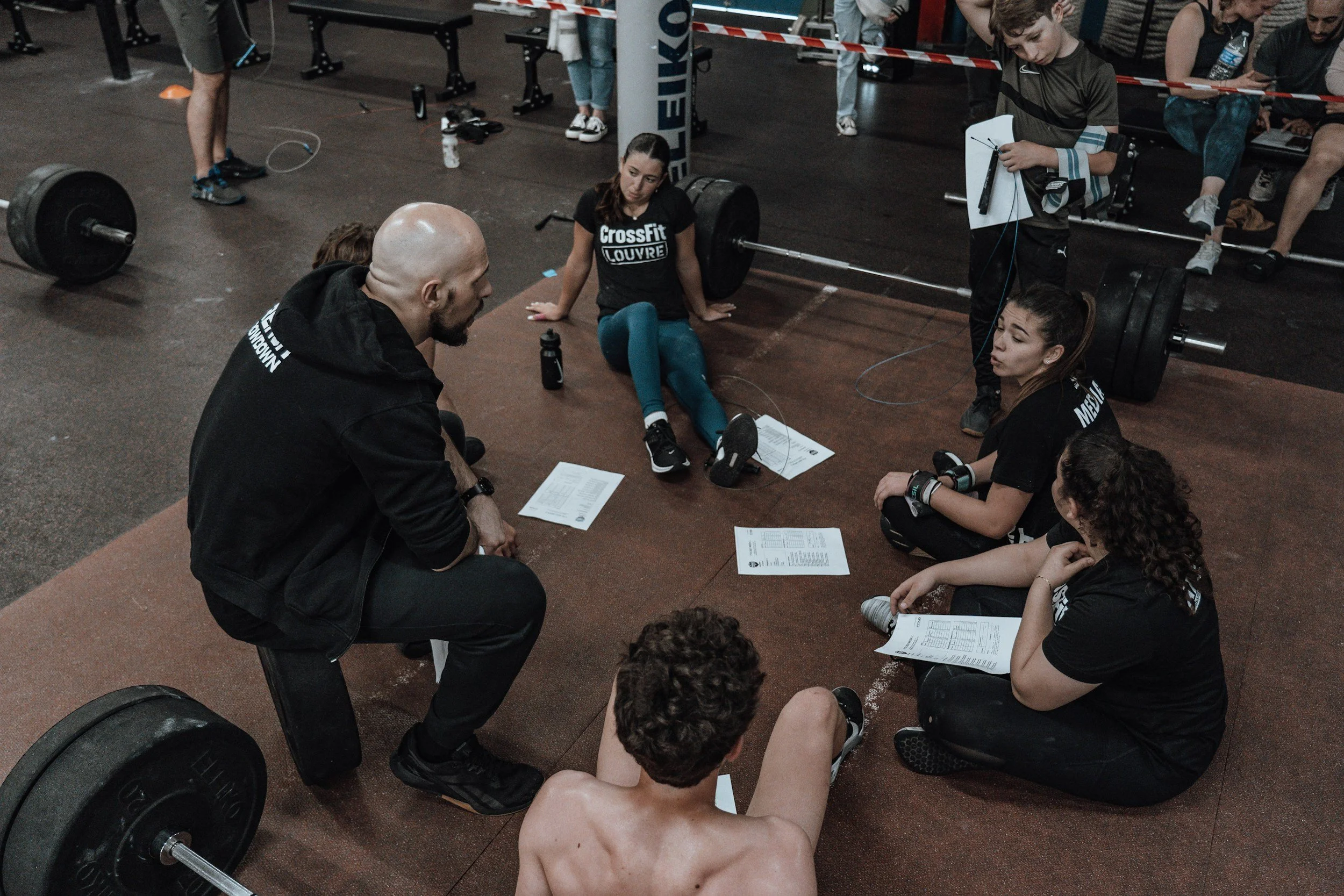 A person lifting weights during a CrossFit workout while others sit on the floor and listen, with gym equipment and a crowd in the background.