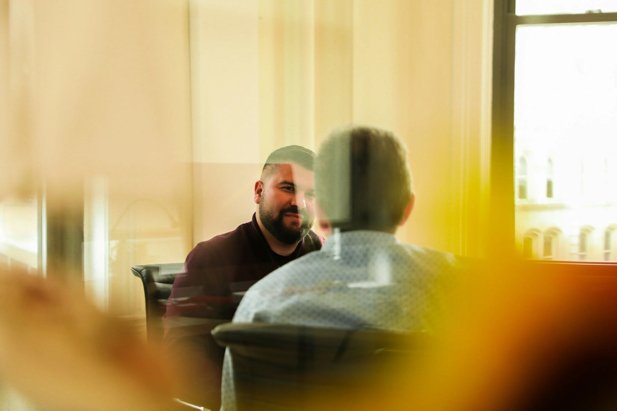 Two men sitting across from each other in a room, seen through a reflective glass or window, with the man on the left smiling and the back of the head of the man on the right visible.