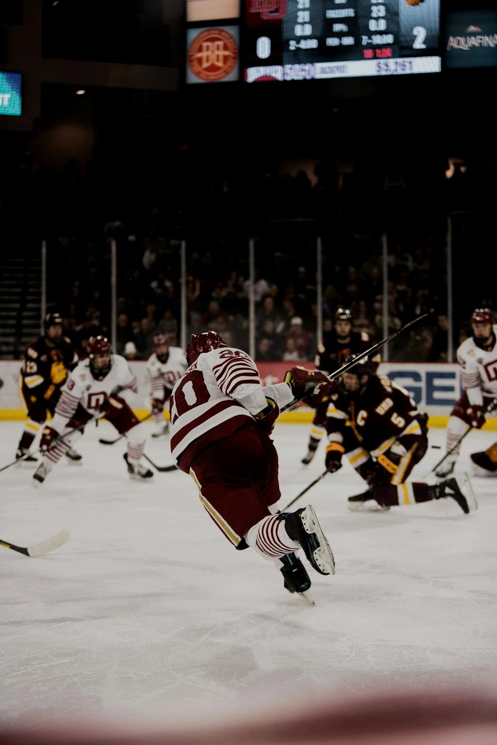 An ice hockey game in progress with players from two teams. One player is in focus, wearing a maroon and white jersey, skating and holding a hockey stick. Opposing players in black and yellow jerseys are seen in the background, with some kneeling or falling on the ice. The arena is filled with spectators, and an electronic scoreboard is visible above.