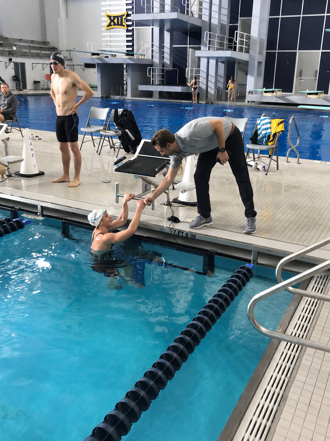 Swimmer in the pool receiving assistance from a coach during a swim meet at an indoor aquatic center, with another swimmer and officials in the background.