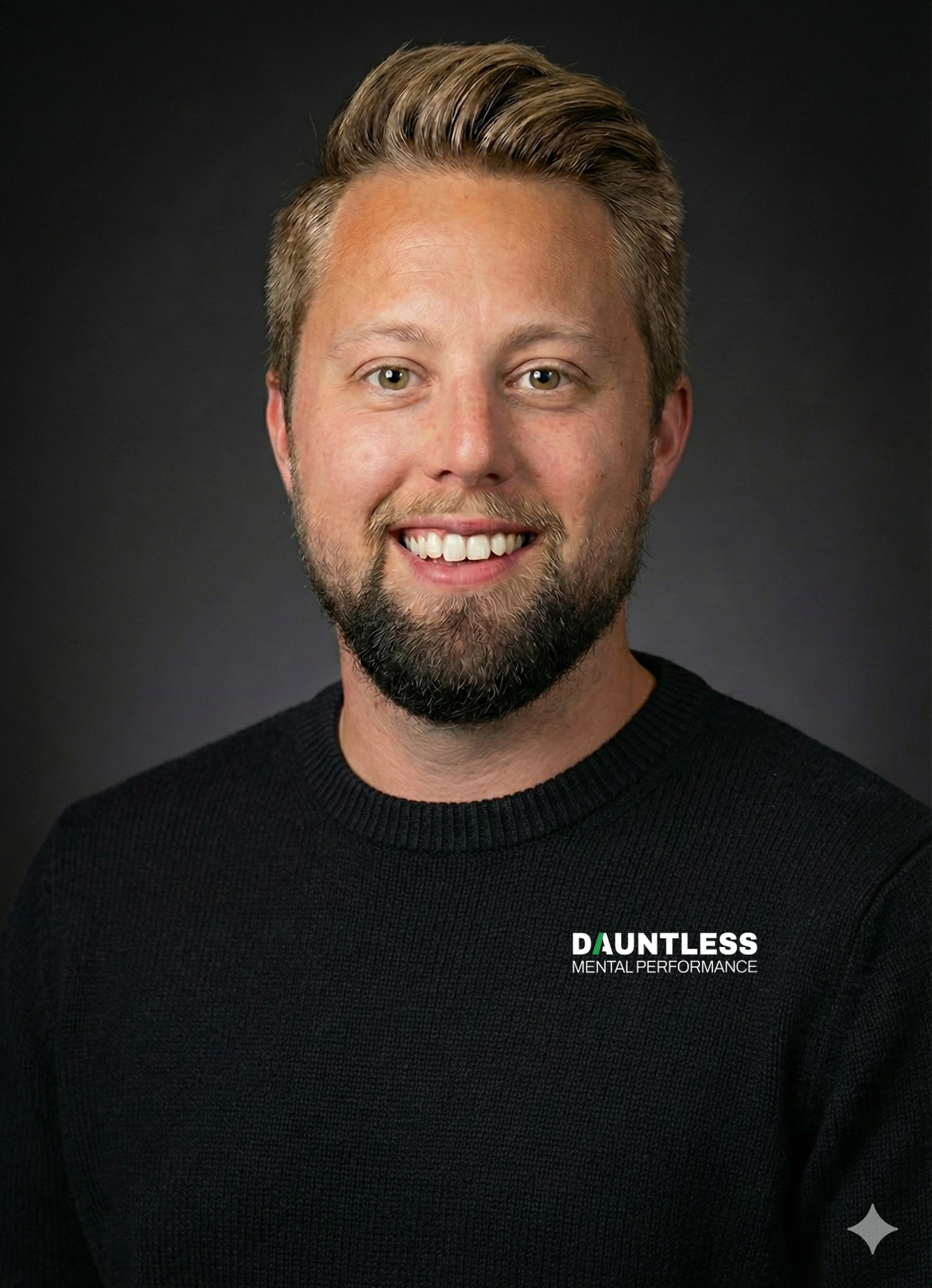 A smiling man with light brown hair, a beard, and light-colored eyes, wearing a black sweater with the text 'DAUNTLESS MENTAL PERFORMANCE' on it, against a dark background.