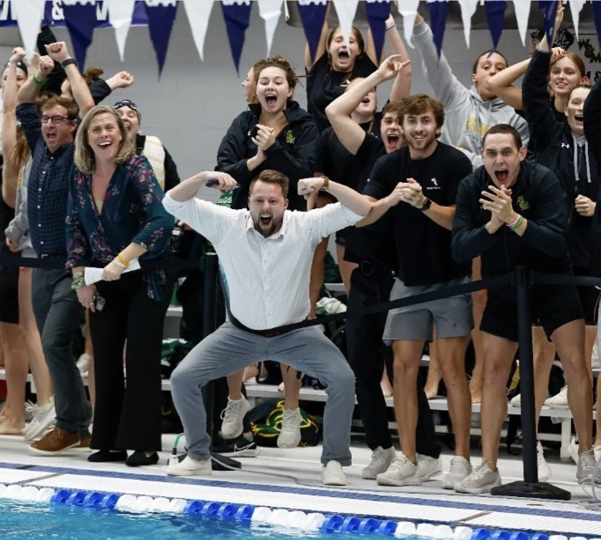 Group of enthusiastic swim team members and coach celebrating beside a swimming pool during a competition, with some individuals flexing and expressing excitement.