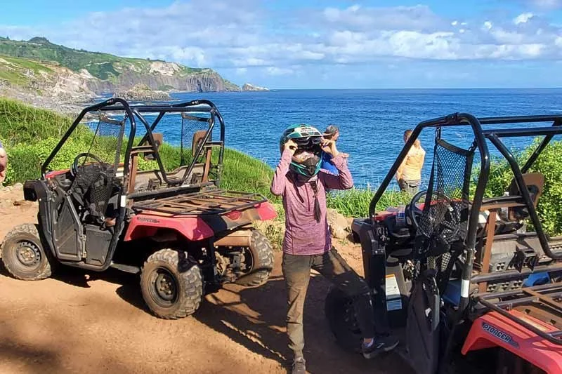 Person adjusting goggles on their head near off-road vehicles by the ocean with cliffs in the background.