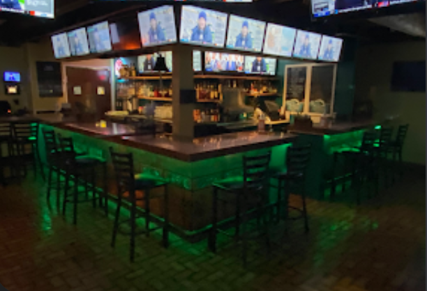 A bar with a U-shaped counter illuminated by green lighting, surrounded by black chairs, with multiple TV screens above showing a sports game or show, and shelves with bottles and glasses behind the counter.