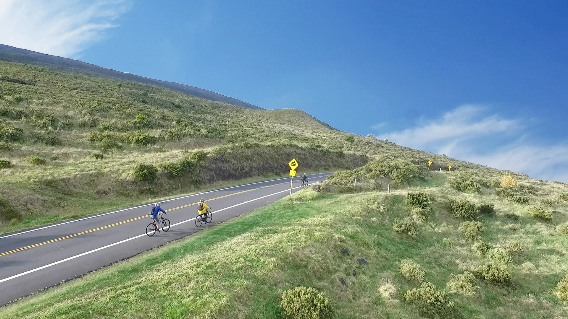 Three cyclists riding on a winding mountain road, surrounded by green hills and a blue sky.