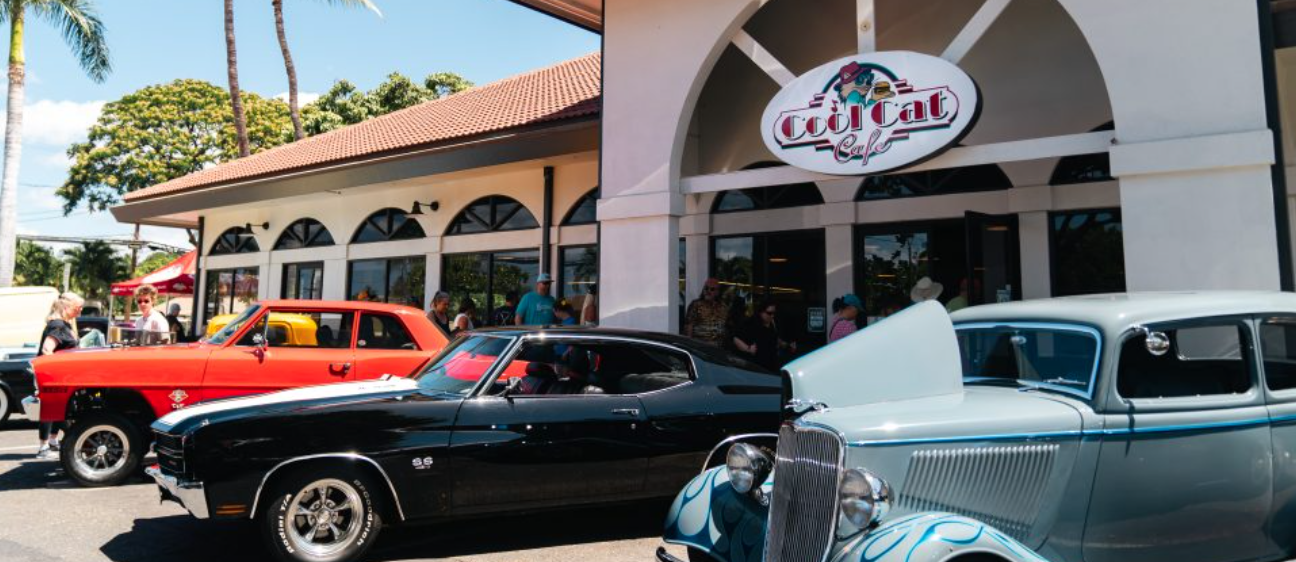A classic car show outside a café named 'Cool Cat Café' with vintage cars parked in front and people walking around.
