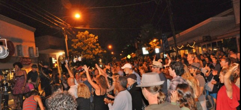 Crowd of people gathered outdoors at night for a street event or concert, some dancing and enjoying live music on a small stage, with streetlights illuminating the scene.