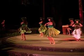 Four hula dancers performing on stage with two seated musicians, in brightly colored costumes, during a nighttime outdoor event.