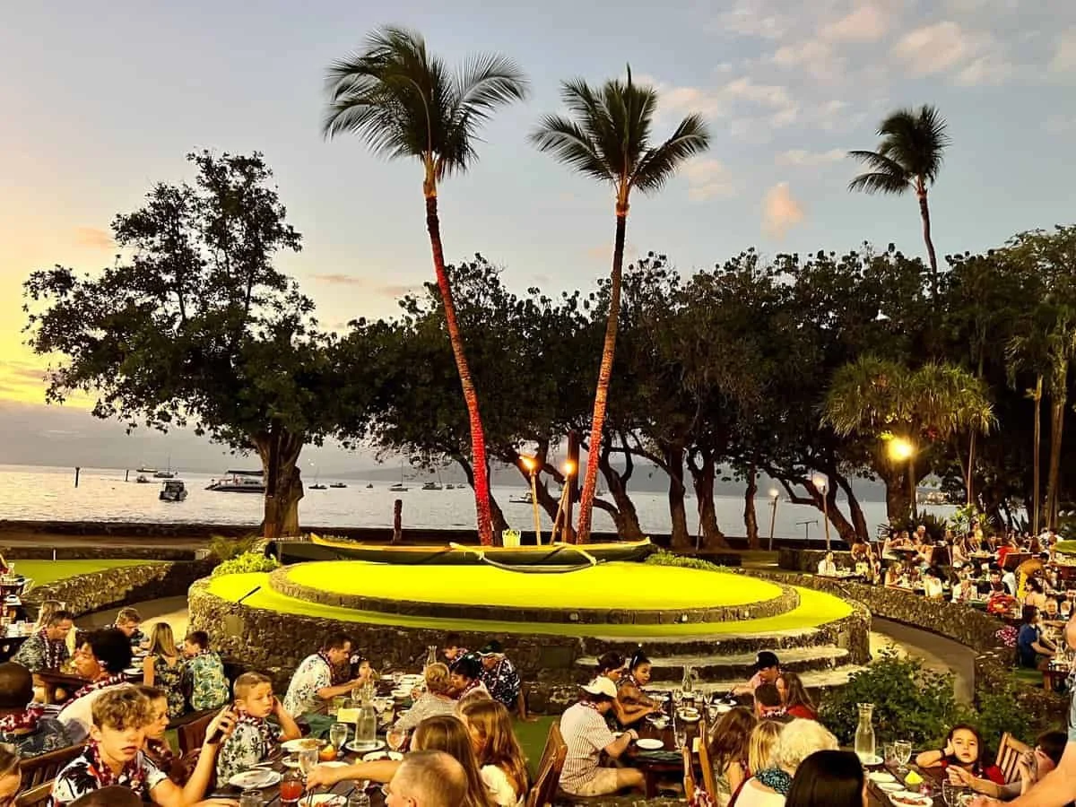 People dining outdoors at a restaurant near a waterfront with palm trees, a grassy area, and boats on the water at sunset.