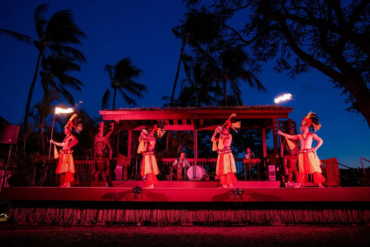 Performers dancing with fire torches on an outdoor stage at night, surrounded by palm trees and tropical scenery.