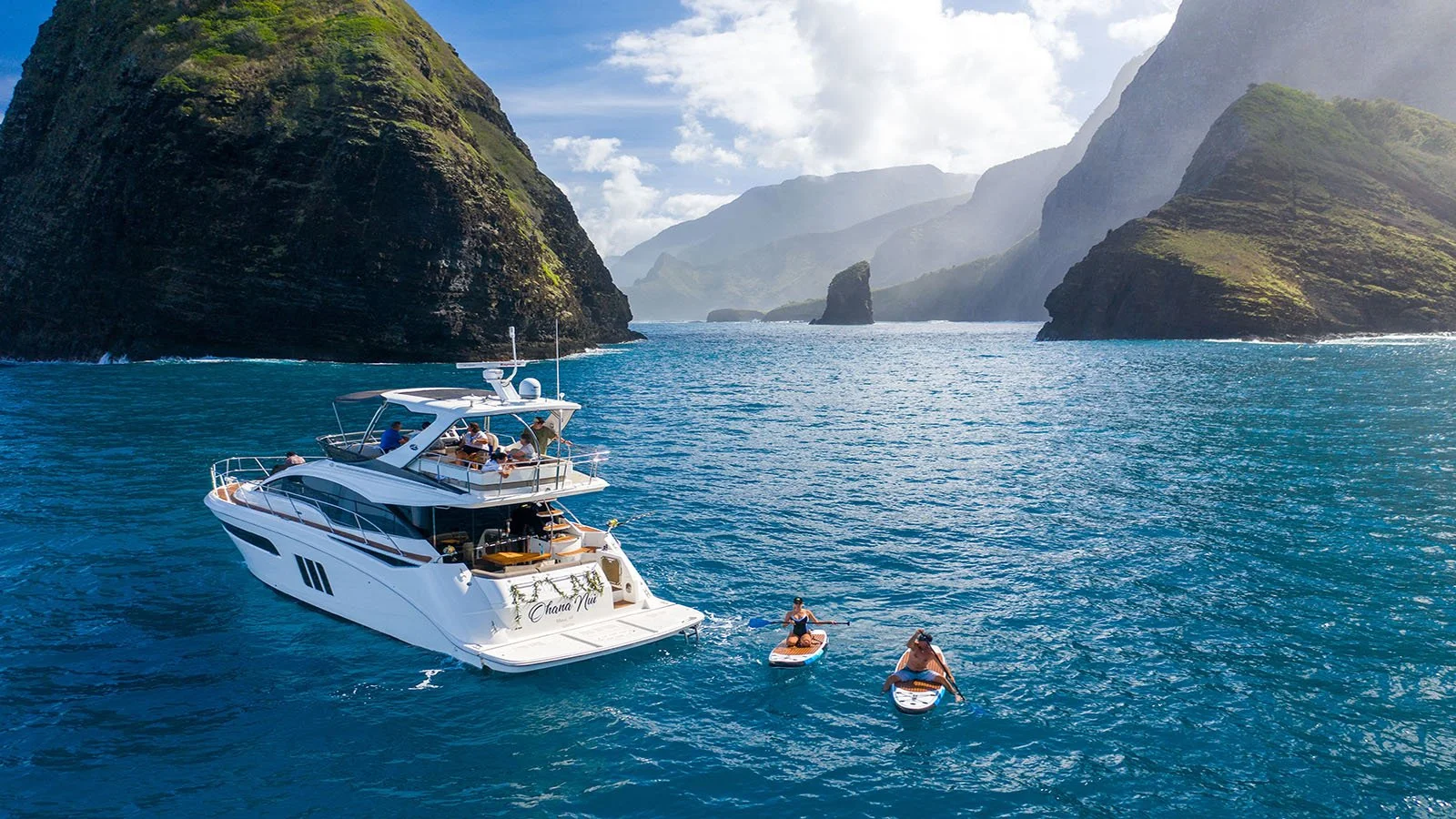 A white yacht with multiple levels on a blue ocean, surrounded by cliffs and mountains under a partly cloudy sky. Two people are paddleboarding near the yacht.