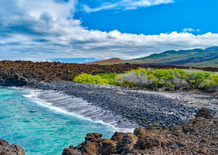 Beach with black volcanic rocks, turquoise water, green trees, and rolling hills under a partly cloudy sky.