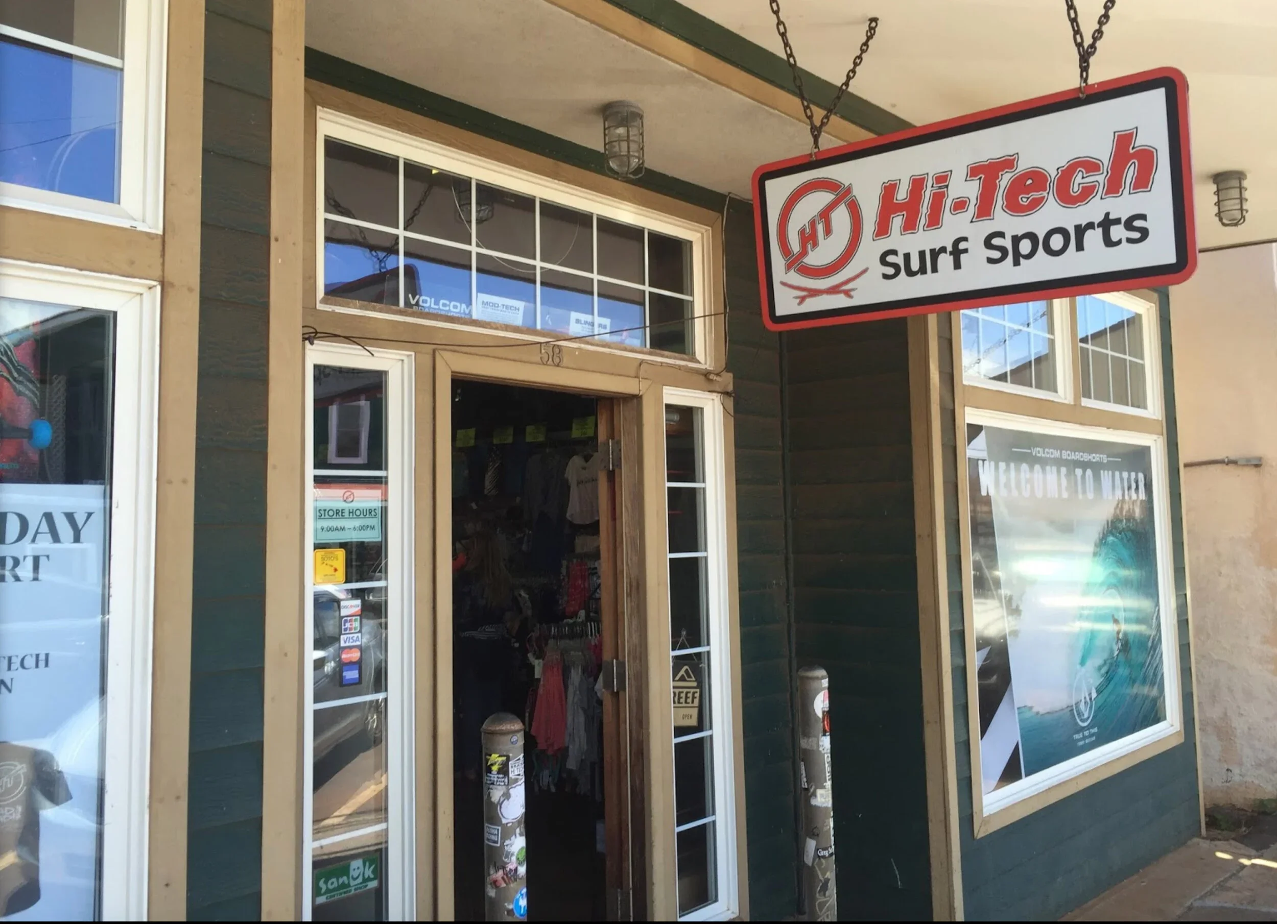 Exterior of a surf sports shop called Hi-Tech Surf Sports with a hanging sign, large window displays, and a door. The shop's store hours are posted on the door.