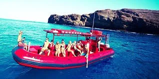 A group of people on a red inflatable boat with a cabin, floating on water near a rocky coastline under a blue sky.