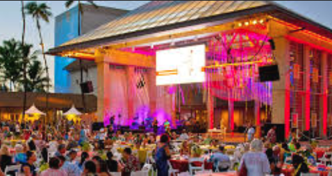 An outdoor concert with a large crowd, stage with pink lighting, and a building with a metal roof in the background.