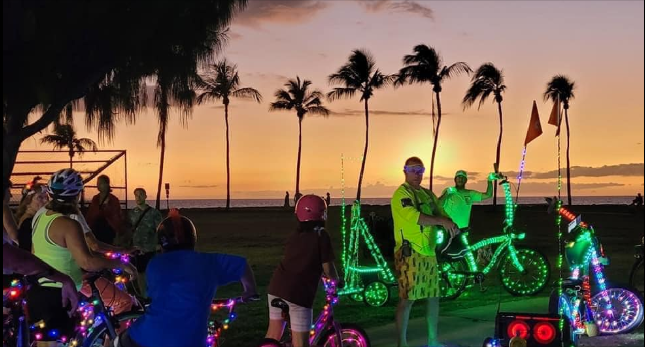 People riding bikes decorated with colorful lights on a beach at sunset, with palm trees in the background.