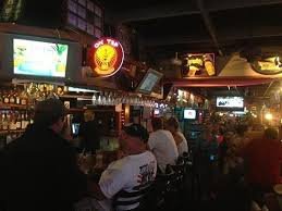 Interior of a bar with patrons sitting at the counter, multiple televisions showing sports, and neon signs, including a prominent Bud Light sign.