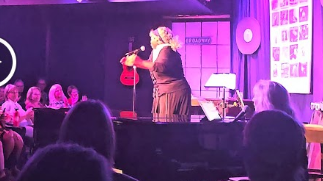 A woman on stage performing with a small guitar, engaging an audience in a cozy, purple-hued venue. The audience is seated, watching attentively.