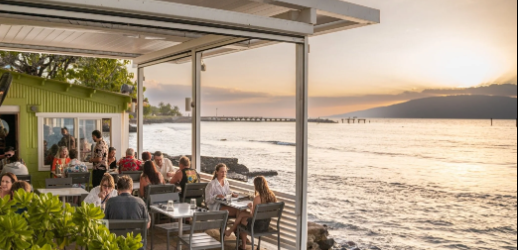 People dining at an outdoor restaurant by the water during sunset.