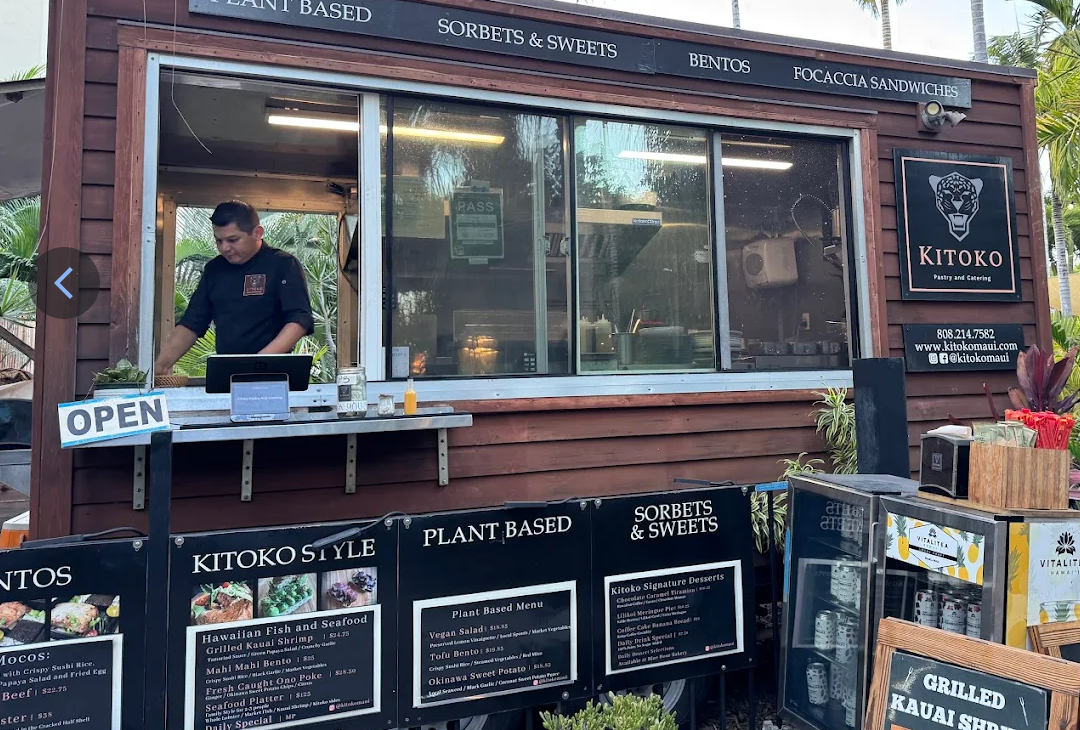 A small restaurant stand with a chef preparing food inside. The stand has signs advertising plant-based, sorbets, sweets, bentos, and sandwiches, and an 'OPEN' sign. The menu features Hawaiian fish, seafood, vegan salads, tofu bento, and sweet potato dishes.