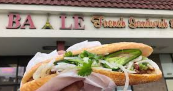 Close-up of a sandwich with lettuce, onion, and other ingredients, held up in front of a French sandwich shop sign.