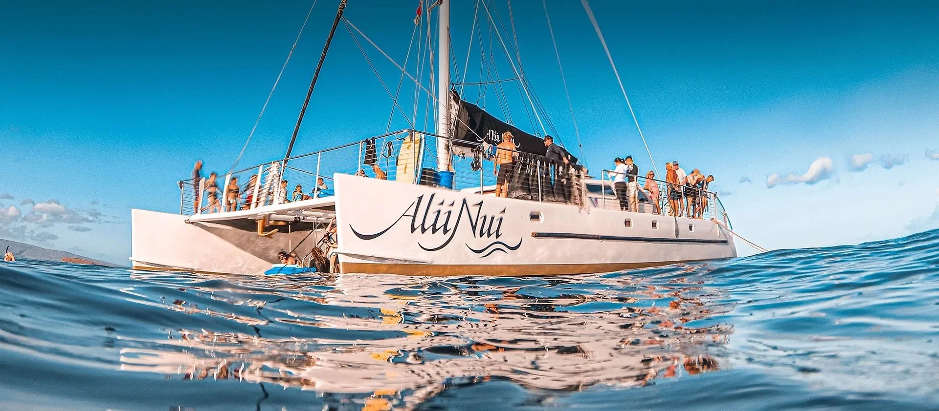 A large white sailing yacht named 'Alii Nui' with people on the deck, floating on calm ocean waters under a blue sky.