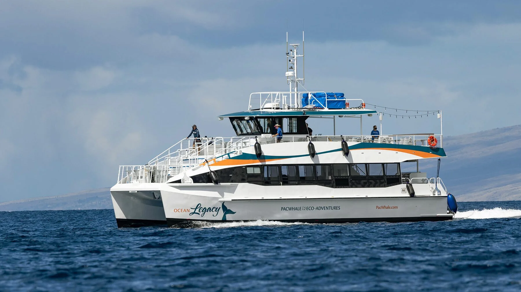 White and teal boat named Ocean Legacy sailing on ocean water with land in the background, with several people on deck