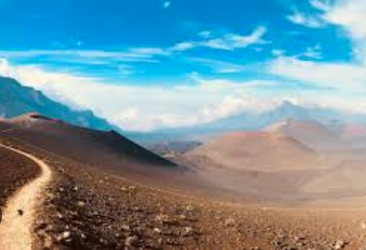 A rugged mountain trail winding through an arid landscape with rolling hills and distant mountains under a cloudy sky.