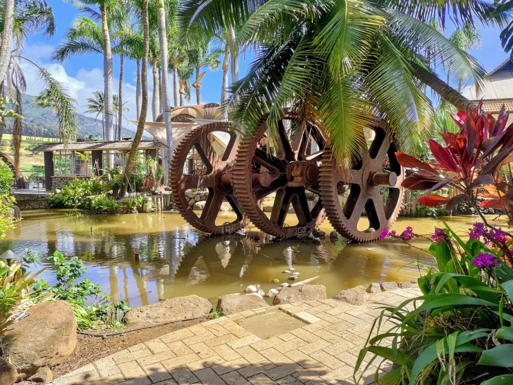Rusty large gear wheels placed in water surrounded by tropical plants and palm trees, with a building and mountains in the background.