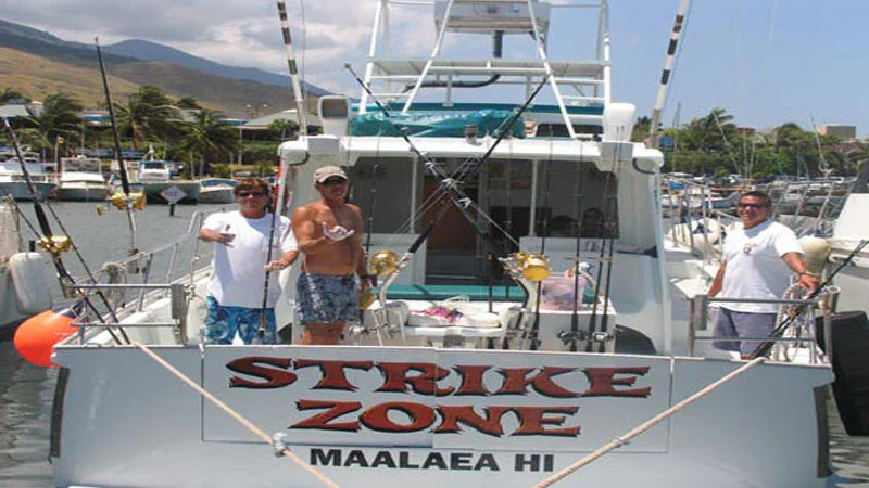 Three men on a boat named 'Strike Zone' at a marina, with mountains and other boats in the background, holding drinks and smiling.