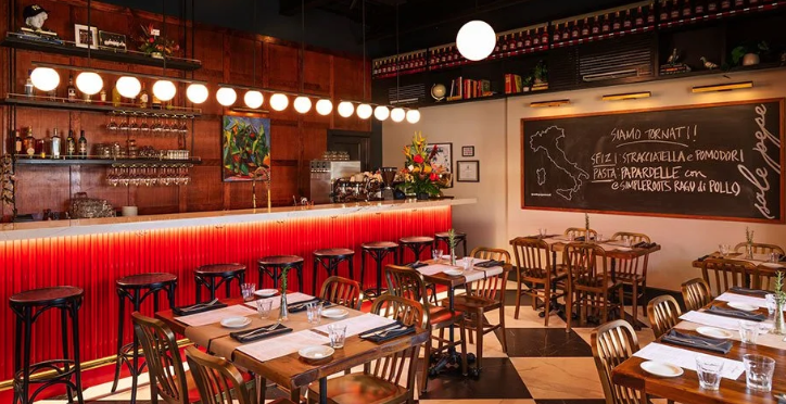 Interior of a cozy restaurant with a bar area, wooden tables and chairs set for dining, a chalkboard menu on the wall, and warm lighting.