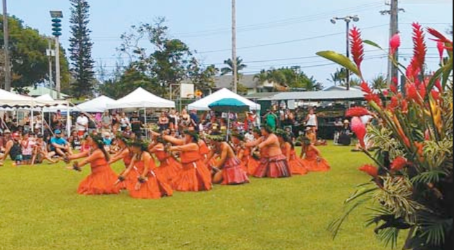 Group of women dancing in orange skirts at an outdoor cultural event with tents and spectators in the background.